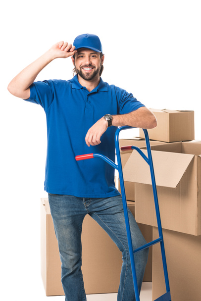 handsome delivery man standing near cardboard boxes and smiling at camera isolated on white  - Φωτογραφία, εικόνα