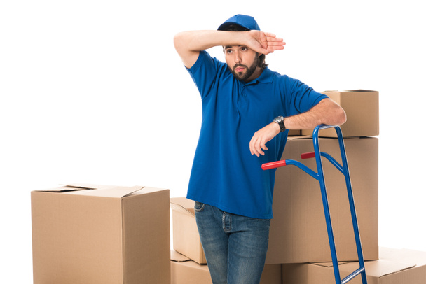 tired delivery man standing between cardboard boxes and looking away isolated on white  - Foto, Imagen