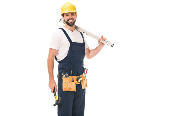 guapo trabajador feliz sosteniendo herramienta de nivel y martillo, sonriendo a la cámara aislada en blanco
 - Foto, Imagen