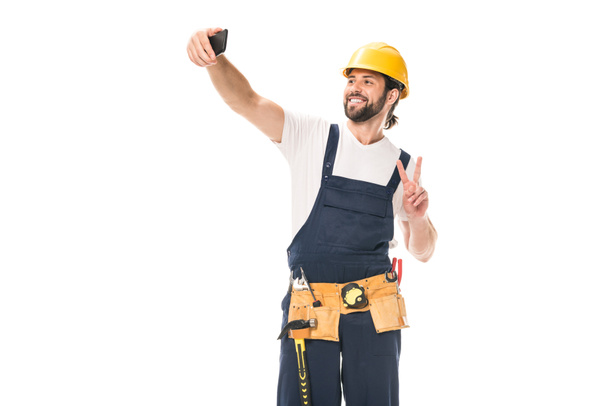 trabalhador bonito feliz em chapéu duro e cinto de ferramentas tirando selfie com smartphone isolado no branco
 - Foto, Imagem