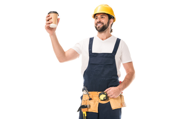 knappe gelukkig workman in gereedschap gordel holding papier beker met koffie te gaan geïsoleerd op wit - Foto, afbeelding