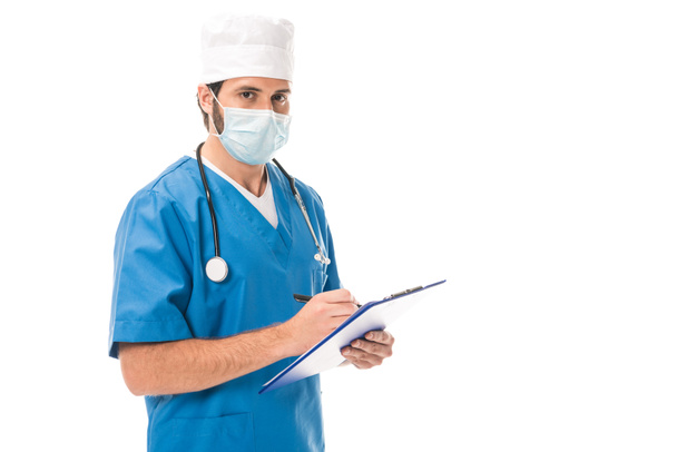 doctor in medical mask writing on clipboard and looking at camera isolated on white - Zdjęcie, obraz