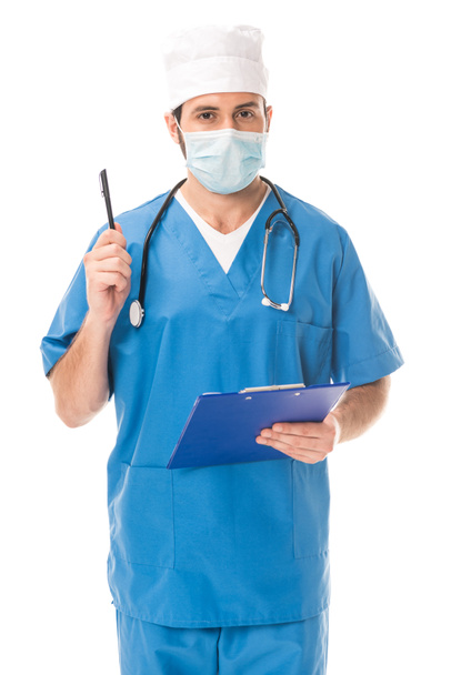 doctor in medical mask holding clipboard with pen and looking at camera isolated on white - Foto, immagini