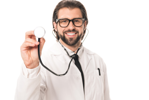 happy bearded doctor in white coat and eyeglasses holding stethoscope and smiling at camera isolated on white - Photo, image