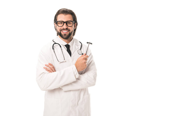 handsome bearded doctor in white coat and eyeglasses holding reflex hammer and smiling at camera isolated on white - Fotó, kép