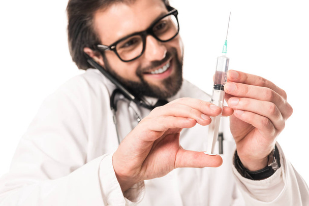smiling doctor holding syringe and talking by smartphone isolated on white  - Foto, Imagem