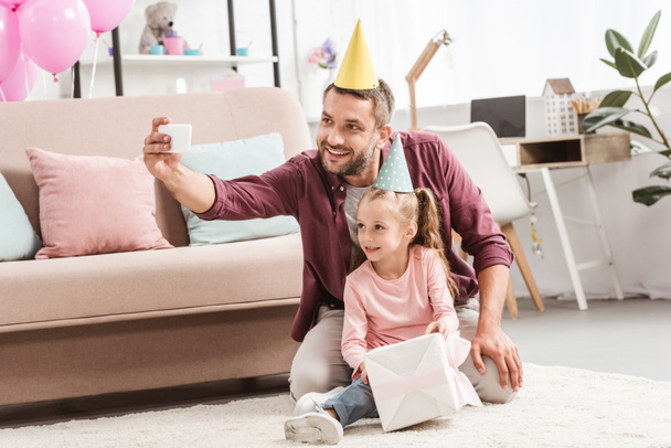 sonriente padre e hija en partido sombreros tomando selfie con regalo para cumpleaños
 - Foto, Imagen