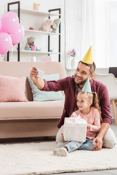 father and adorable daughter in party hats taking selfie with gift for birthday - Foto, Imagem