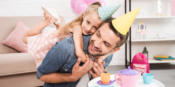 father and daughter in cone hats piggybacking while playing tea party at home - Photo, Image