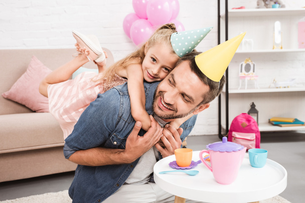 padre piggybacking hija en cono sombrero mientras jugando té fiesta en casa
 - Foto, imagen