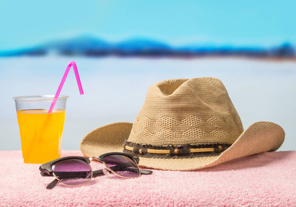 Διακοπές περιπέτειας και το καλοκαίρι διακοπές έννοια με τον απαραίτητο εξοπλισμό. Γείσο Καπέλο, γυαλιά ηλίου και κίτρινο γευστικό ρόφημα σε πετσέτα στην παραλία. Αναψυχής σε όμορφο και ηλιόλουστο τροπικό παράδεισο. - Φωτογραφία, εικόνα
