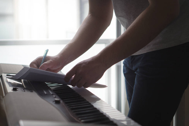 Komponist und Songwriter, der Noten oder Texte auf Papier am Piano schreibt. Musiker schreiben einen Song im heimischen Studio auf einem digitalen Instrument. Aufgeregt über neue Idee oder Melodie. Schattenhafte dunkle Umgebungsbeleuchtung. - Foto, Bild