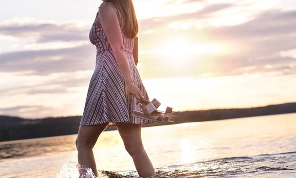 Έννοια της ελευθερίας και της ευτυχίας. Ελκυστική κοπέλα περπάτημα στο νερό της λίμνης στο ηλιοβασίλεμα. Ανέμελη και χαρούμενος τρόπος ζωής. Κρατώντας στο χέρι σανδάλια. Καυτή νύχτα του καλοκαιριού. - Φωτογραφία, εικόνα