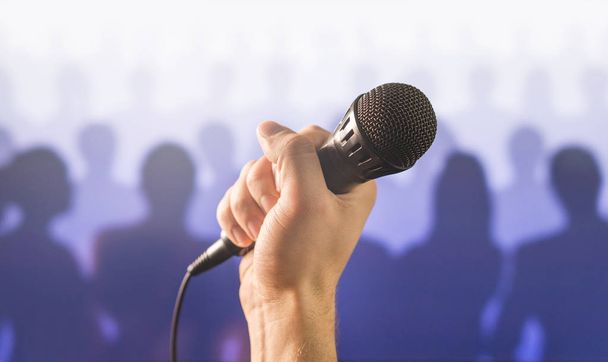 Falar em público e dar conceito de discurso. Mão segurando microfone na frente de um público silhueta e multidão de pessoas. Cantando ao microfone em karaoke ou conceito de show de talentos
. - Foto, Imagem