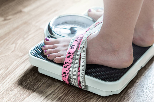 Feet tied up with measuring tape to a weight scale. Addiction and obsession to weight loss. Anorexia and eating disorder concept. - Photo, Image