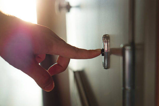 Finger pressing doorbell in sunny apartment building corridor. Close up of male hand ringing door bell in a block of flats. Salesman, fundraiser, guest or visitor behind door. - Фото, изображение