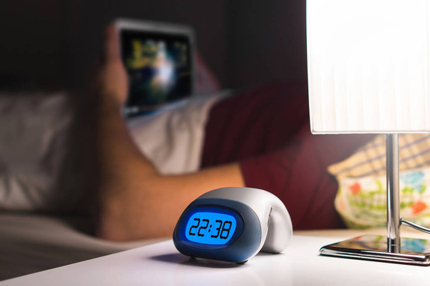 Az ember használ tabletta, vagy néz film vagy sorozat patak online az ágyban, mielőtt elalszik. A hálószobában éjjeliszekrényre digitális ébresztőóra. - Fotó, kép