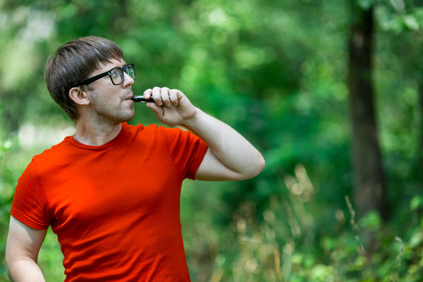 A young white bearded man in red t-shirt and in glasses is vaping an electronic cigarette in the forest in the summer. Close up. - Foto, Bild