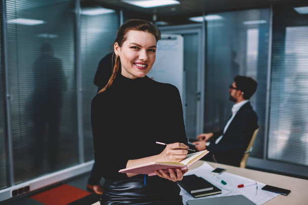 Portrait of cute beautiful young business woman smiling  in office with colleagues on background, woman notes ideas for profitable project and looking at camera attractive female secretary on table - Foto, afbeelding