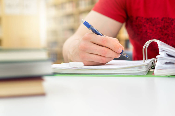 Young athletic man and student studying and writing notes in public or school library in college or university. Stack and pile of books, pen and paper on table. - Photo, Image