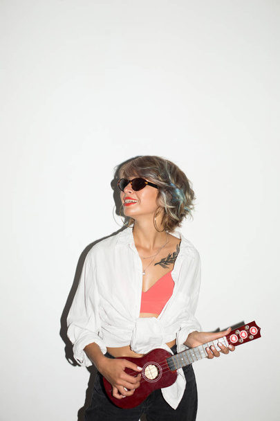 Cool fille souriante en chemise blanche et lunettes de soleil regardant joyeusement de côté en jouant sur la petite guitare sur fond blanc
  - Photo, image