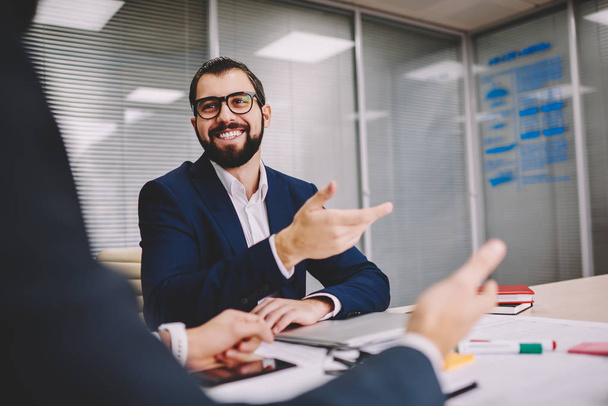 Happy bearded proud ceo in eyeglasses for vision correction communicating with business colleague about productive solution in office interior.Prosperous male executive manager dressed in formal wear - 写真・画像