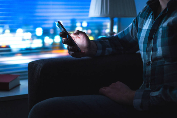 Man using mobile phone home late at night. Guy looking messages with modern cellphone in dark. Unfaithful husband cheating and texting with another woman. Secret or illegal business. Sitting on couch. - Foto, imagen