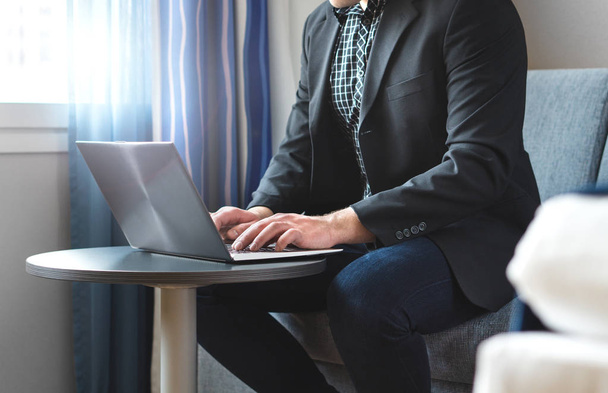 Obchodní muž pracující s přenosným počítačem v hotelovém pokoji. Podnikatel dělá vzdálenou práci s počítačem. Člověk sedí a psaní. Výkonný psaní firemního e-mailu na služební cestě. - Fotografie, Obrázek