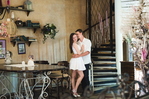 Young couple embracing in a cafe. A long legged shy girl smiling in her man's hands, wooden dark furniture around them - Foto, imagen