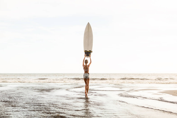Young surfer girl in sexy white bikini and short denim shorts stay in river and pose with surf short board. Modern family lifestyle, people water sport adventure camp, extreme swim on summer vacation - 写真・画像