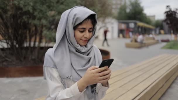 Young woman in hijab texting on a bench in a city - Metraje, vídeo