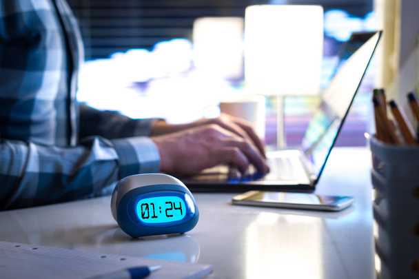 Man working late. Workaholic or being behind schedule concept. Business person in modern office building or home at night using laptop. Time in digital clock on table in workstation. - Photo, Image