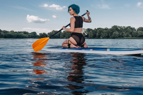athletic tattooed girl sitting on paddle board on river - Photo, Image
