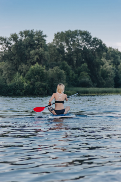 back view of blonde girl sitting on paddle board on river - Photo, Image
