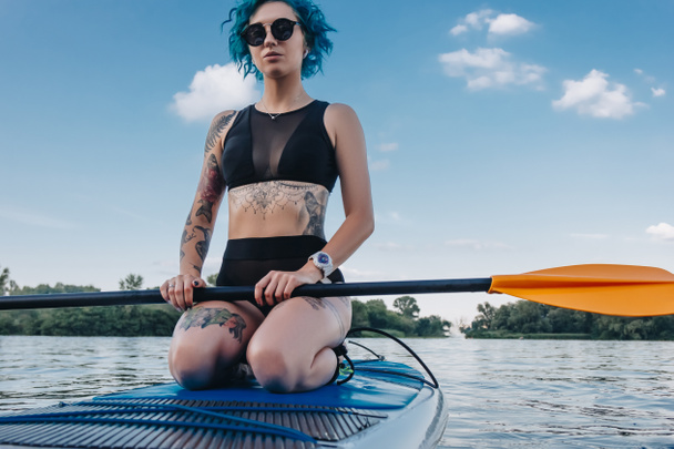attractive tattooed girl in sunglasses sitting on paddle board on river - Фото, изображение