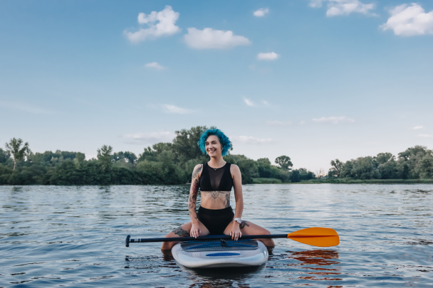 beautiful tattooed girl with blue hair sitting on sup board on river - Foto, Imagem