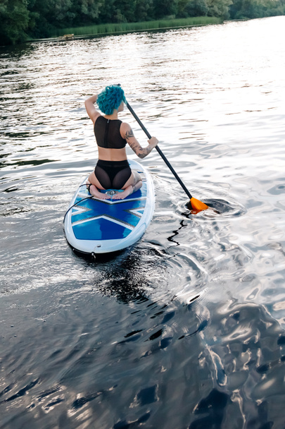 back view of sportive girl with blue hair paddle surfing on river - Photo, Image