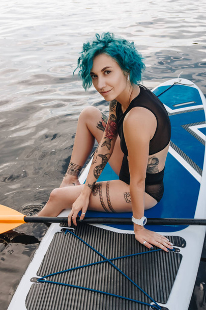 attractive athletic girl with blue hair resting on paddle board on river - Zdjęcie, obraz