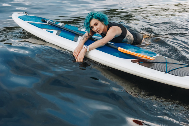 kaunis urheilullinen tatuoitu nuori nainen, jolla on siniset hiukset rentouttava sup aluksella vedessä
 - Valokuva, kuva