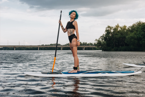 beautiful dreamy athletic tattooed woman standup paddleboarding on river - Foto, Bild