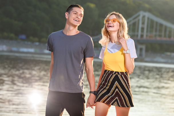 laughing young couple holding hands and walking on river beach in evening - Foto, afbeelding