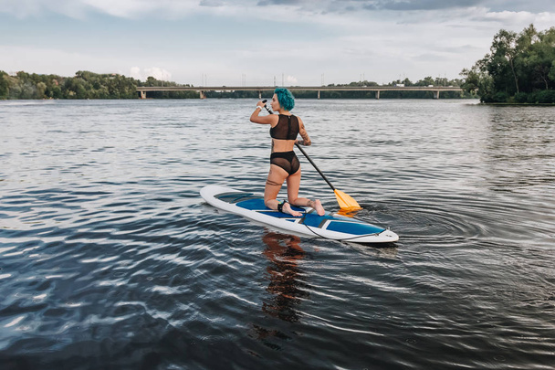 mooie sportieve getatoeëerd vrouw in bikini peddel aan boord op de rivier - Foto, afbeelding