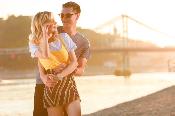 Парень обнимает подружку на пляже на закате, она касается солнцезащитных очков
 - Фото, изображение