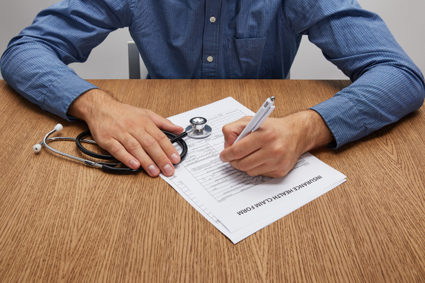 colpo ritagliato di persona che tiene lo stetoscopio e firma modulo di assicurazione al tavolo di legno
  - Foto, immagini