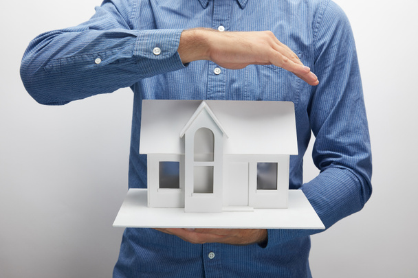 Кадр, на котором мужчина держит в руках модель маленького дома на сером страховом покрытии
 - Фото, изображение