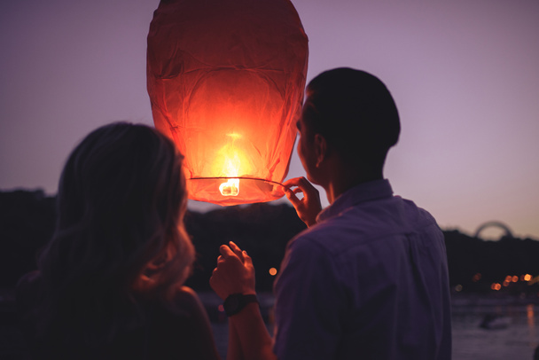 rear view of young couple launching sky lantern on river beach in evening - Photo, Image