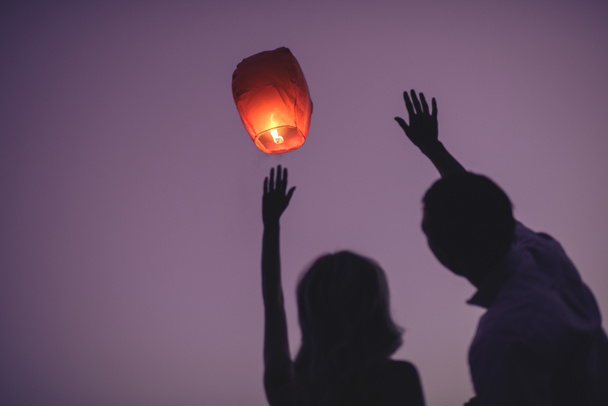 silhouettes of couple waving hands to flying chinese lantern in violet evening sky - Photo, Image