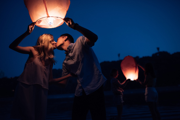 jong koppel kussen en lancering van hemel lantaarn op rivier strand in avond - Foto, afbeelding