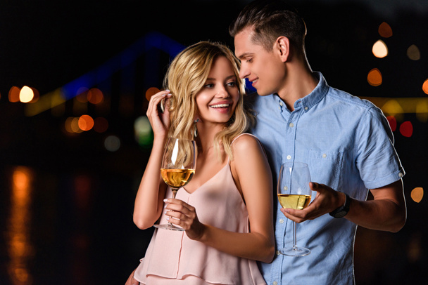happy young couple standing with glasses of white wine on river beach in evening - Foto, afbeelding