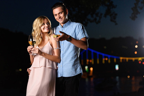 χαμογελώντας νεαρό ζευγάρι στέκεται με ποτήρια κρασιού στην παραλία ποτάμι στο βράδυ - Φωτογραφία, εικόνα